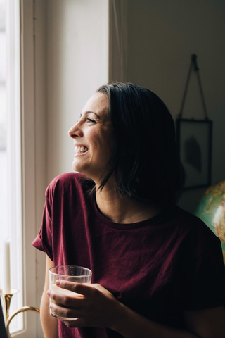 Nachdenklich lächelnde Frau trinkt Tee und schaut durch ein Fenster, lizenzfreies Stockfoto