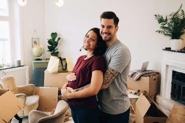 Smiling man embracing pregnant woman while standing against boxes in living room - MASF00180