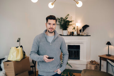 Portrait of smiling man holding mobile phone while standing with hand in pocket at home - MASF00187