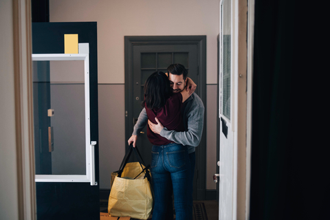 Frau umarmt Mann mit Tasche, während sie im Korridor steht, gesehen vom Eingang aus, lizenzfreies Stockfoto