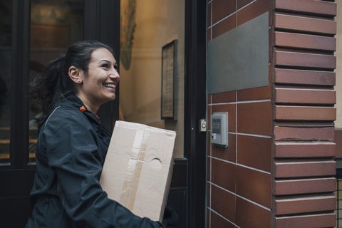 Lächelnder weiblicher Bote, der wegschaut, während er eine Kiste durch eine geschlossene Tür trägt, lizenzfreies Stockfoto