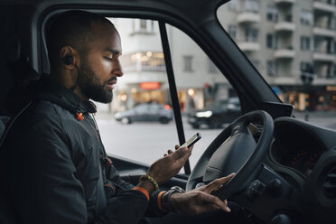 Side view of male driver looking at mobile phone while sitting in delivery van at city - MASF00258