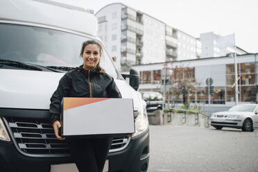 Porträt einer lächelnden Arbeiterin, die eine Kiste trägt und gegen einen Lieferwagen in der Stadt steht - MASF00269