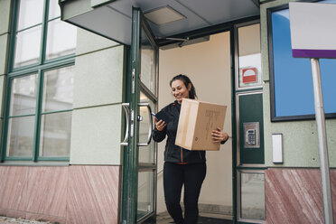 Lächelnde Arbeiterin mit Kiste, die ein Mobiltelefon benutzt, während sie aus dem Gebäude kommt - MASF00270