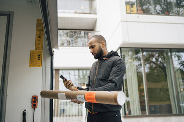 Männlicher Bote mit Paket, der ein Mobiltelefon benutzt, während er am Lieferwagen steht - MASF00276