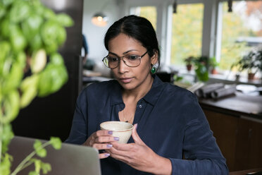 Geschäftsfrau mit Tasse in der Hand und Blick auf den Laptop im Heimbüro - MASF00298