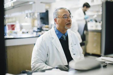 Doctor working at desk while male coworker working in background at hospital - CAVF34480