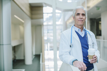 Portrait of male doctor holding disposable cup while standing in hospital corridor - CAVF34513