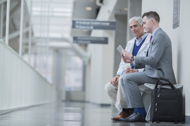 Male doctors discussing over tablet computer while sitting on seat in hospital corridor - CAVF34517