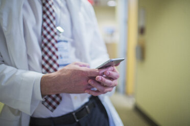 Männlicher Arzt, der ein Mobiltelefon benutzt, während er im Bürokorridor steht - CAVF34566