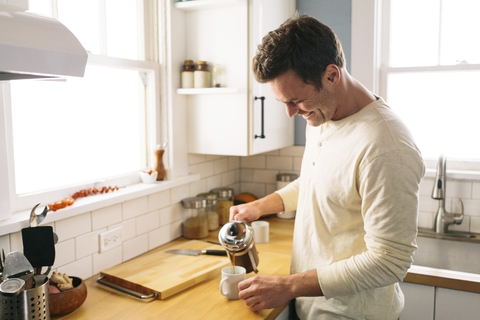 Glücklicher Mann gießt Kaffee ein, während er an der Küchentheke zu Hause steht, lizenzfreies Stockfoto