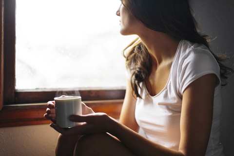 Midsection of Woman with cup sitting by window at home stock photo