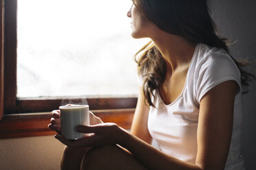 Midsection of Woman with cup sitting by window at home - CAVF34580