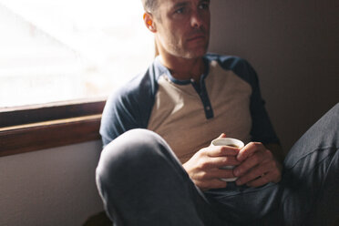 Thoughtful man with coffee cup sitting by window at home - CAVF34582