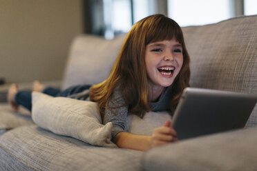 Fröhliches Mädchen, das einen Tablet-Computer benutzt, während es zu Hause auf dem Sofa liegt - CAVF34608
