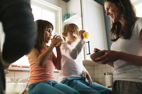 Mutter schaut auf Kinder, die Saft trinken, während sie auf dem Küchentisch sitzen, lizenzfreies Stockfoto