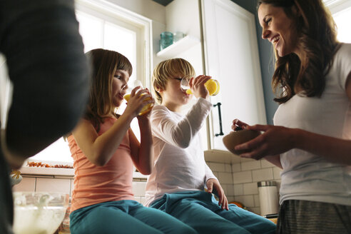 Mutter schaut auf Kinder, die Saft trinken, während sie auf dem Küchentisch sitzen - CAVF34624