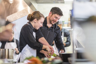 Male chef assisting female coworker in chopping vegetables at restaurant kitchen - CAVF34694