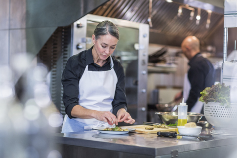 Weibliche Köchin garniert Essen, während ein Mitarbeiter im Hintergrund in der Restaurantküche arbeitet, lizenzfreies Stockfoto