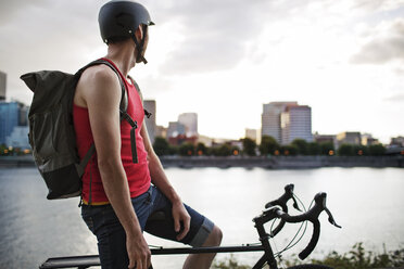 Thoughtful athlete sitting on bicycle by lake in city - CAVF34925