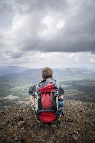 Rückansicht eines Wanderers mit Rucksack auf einem Berg sitzend gegen einen bewölkten Himmel, lizenzfreies Stockfoto