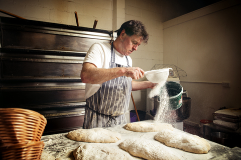 Männlicher Bäcker, der in einer Bäckerei Mehl auf den Brotteig streut, lizenzfreies Stockfoto