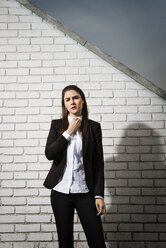 Porträt einer selbstbewussten Geschäftsfrau, die ihren Kragen zurechtrückt, während sie an einer Backsteinmauer steht - CAVF35041