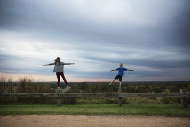 Teenage girl and boy standing on one leg on wooden fence against sky - CAVF35077