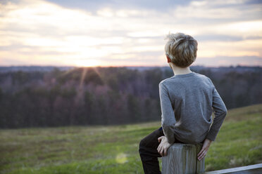 Boy looking at sunset sitting on fence - CAVF35109