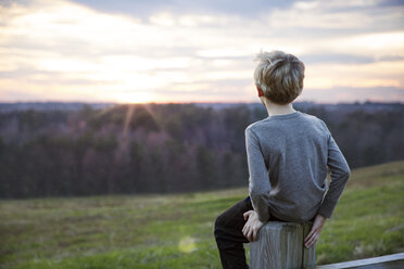 Junge mit Blick auf den Sonnenuntergang auf dem Zaun sitzend - CAVF35109