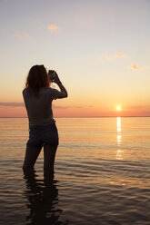 Rückansicht eines jugendlichen Mädchens, das den Sonnenuntergang fotografiert, während es im Meer steht - CAVF35157