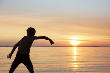 Rückansicht eines Jungen, der bei Sonnenuntergang am Strand Steinchen springt - CAVF35162