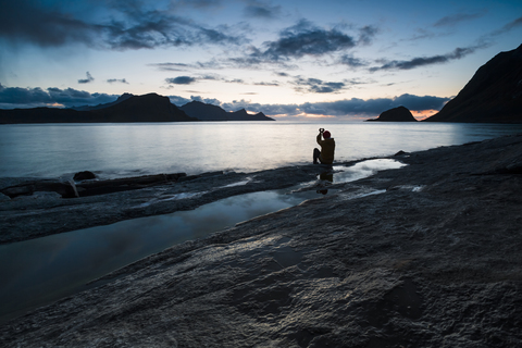 Norwegen, Lofoten-Inseln, Haukland Strand, Mann sitzt auf einem Felsen und baut mit den Fingern ein Herz, lizenzfreies Stockfoto