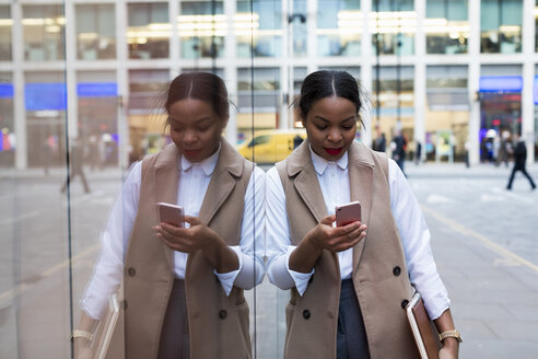 Smiling businesswoman leaning against glass pane looking at cell phone - MAUF01388