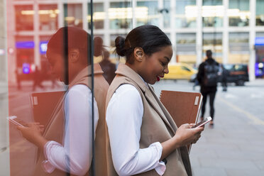 Smiling businesswoman leaning against glass pane using cell phone - MAUF01391