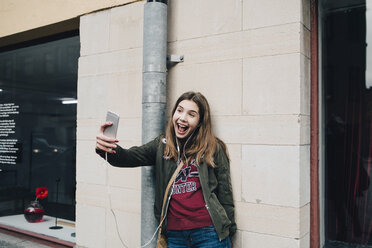 Glückliches Mädchen mit offenem Mund nimmt Selfie durch Smartphone, während gegen die Wand stehen - MASF00370