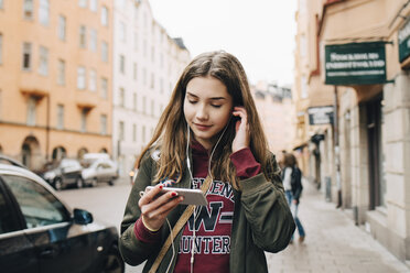 Lächelndes Mädchen hört Musik über ihr Smartphone, während sie auf dem Gehweg in der Stadt spazieren geht - MASF00371