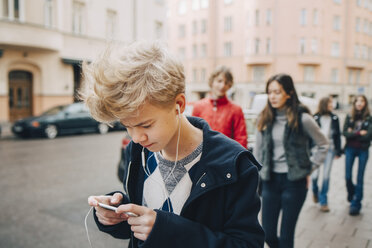 Jugendlicher, der ein Mobiltelefon benutzt, während er mit Freunden auf dem Bürgersteig in der Stadt spazieren geht - MASF00372