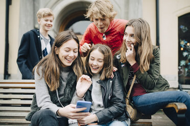 Glückliche Freunde schauen auf ihr Smartphone, während sie auf einer Bank in der Stadt sitzen - MASF00377