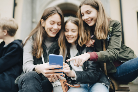 Low angle view of smiling friends looking at smart phone while sitting on bench in city stock photo
