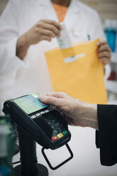 Abgeschnittene Hand einer älteren Kundin, die mit einer Kreditkarte kontaktlos bezahlt, gegenüber einem Apotheker an der Kasse in S - MASF00382