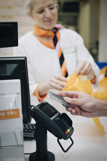 Abgeschnittene Hand eines männlichen Kunden, der eine kontaktlose Zahlung per Kreditkarte gegenüber einem Apotheker an der Kasse eines Geschäfts verwendet - MASF00383