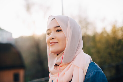 Lächelnde junge muslimische Frau hört Musik über In-Ear-Kopfhörer gegen den klaren Himmel - MASF00394