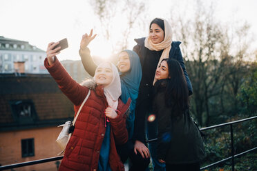 Happy young Muslim woman taking selfie with friends by railing in city - MASF00396