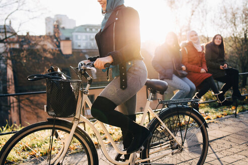 Niedriger Ausschnitt einer Frau auf dem Fahrrad, die von Freundinnen begleitet wird, die auf einem Geländer in der Stadt sitzen - MASF00401