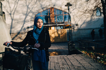 Confident young woman walking with bicycle on footpath against building in city - MASF00402