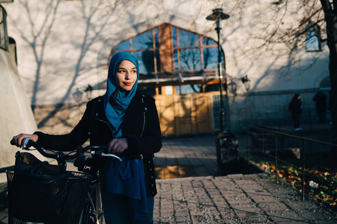 Selbstbewusste junge Frau, die mit dem Fahrrad auf dem Gehweg vor einem Gebäude in der Stadt spazieren geht - MASF00402