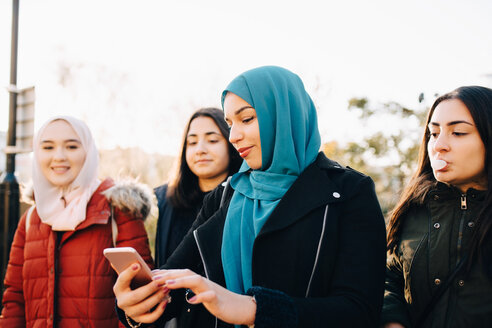 Junge muslimische Frau benutzt ihr Smartphone, während sie mit ihren Freundinnen vor dem Himmel steht - MASF00403