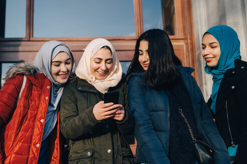 Glückliche muslimische Freundinnen, die sich ein Smartphone teilen, stehen vor einer Eingangstür in der Stadt - MASF00418