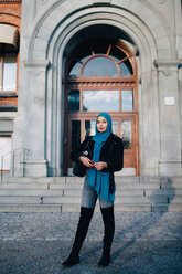 Ganzkörperporträt einer selbstbewussten jungen muslimischen Frau, die mit ihrem Smartphone vor der Eingangstür steht - MASF00420